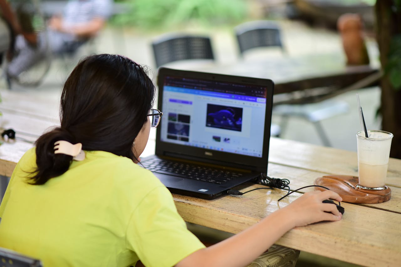 Ask Proctor - A Woman using a laptop in an outdoor café setting with a beverage nearby, focused on work.